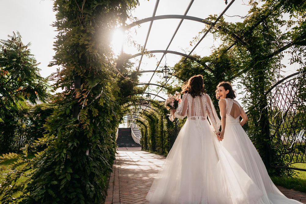 Two women getting married under a pergola with plants and sunshine, celebrating their love and commitment to each other.